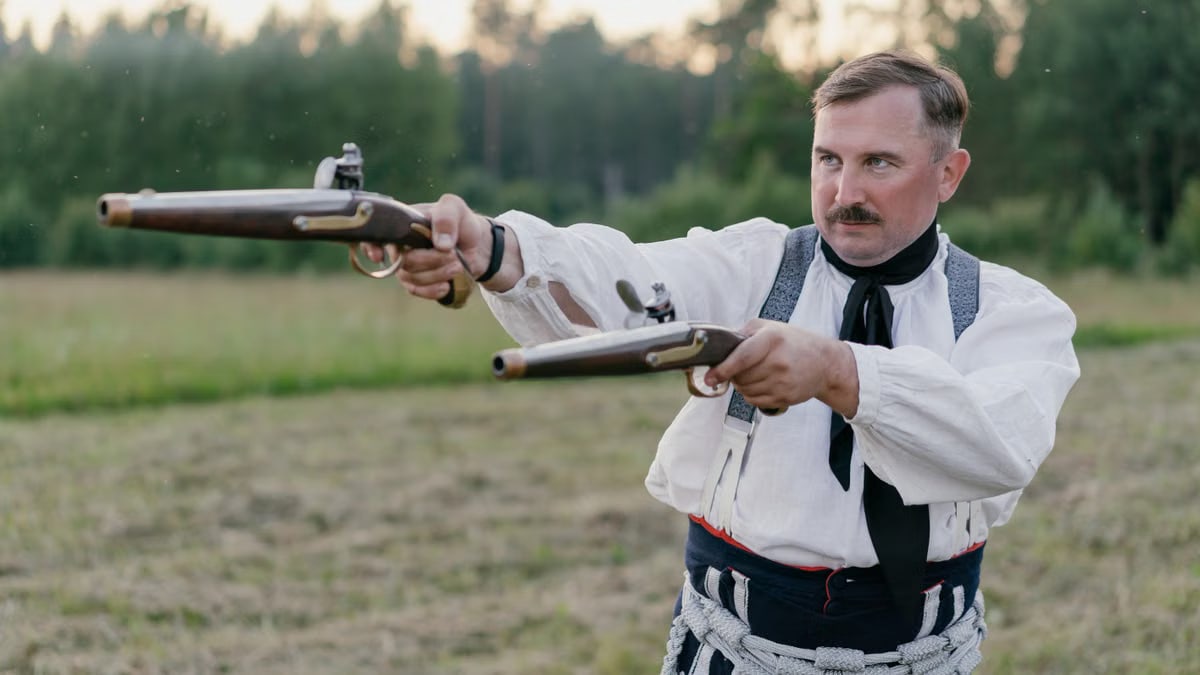 Man in traditional attire aims replica flintlock pistols outdoors.